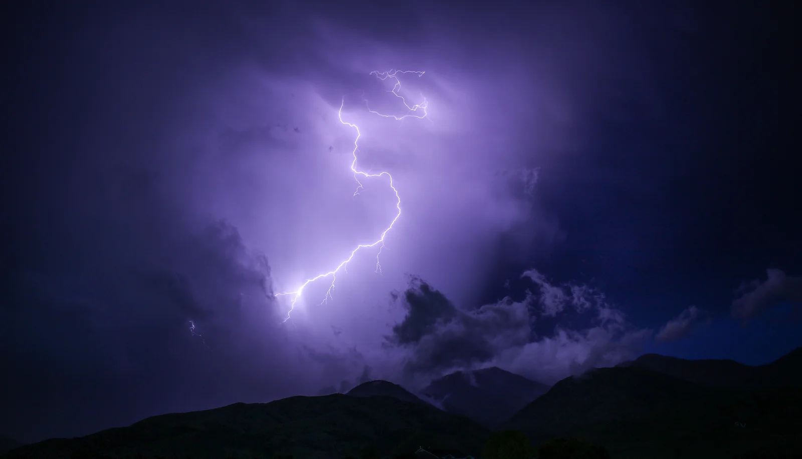 Heller Blitz zwischen dunklen Gewitterwolken über einer Bergkette bei Nacht.