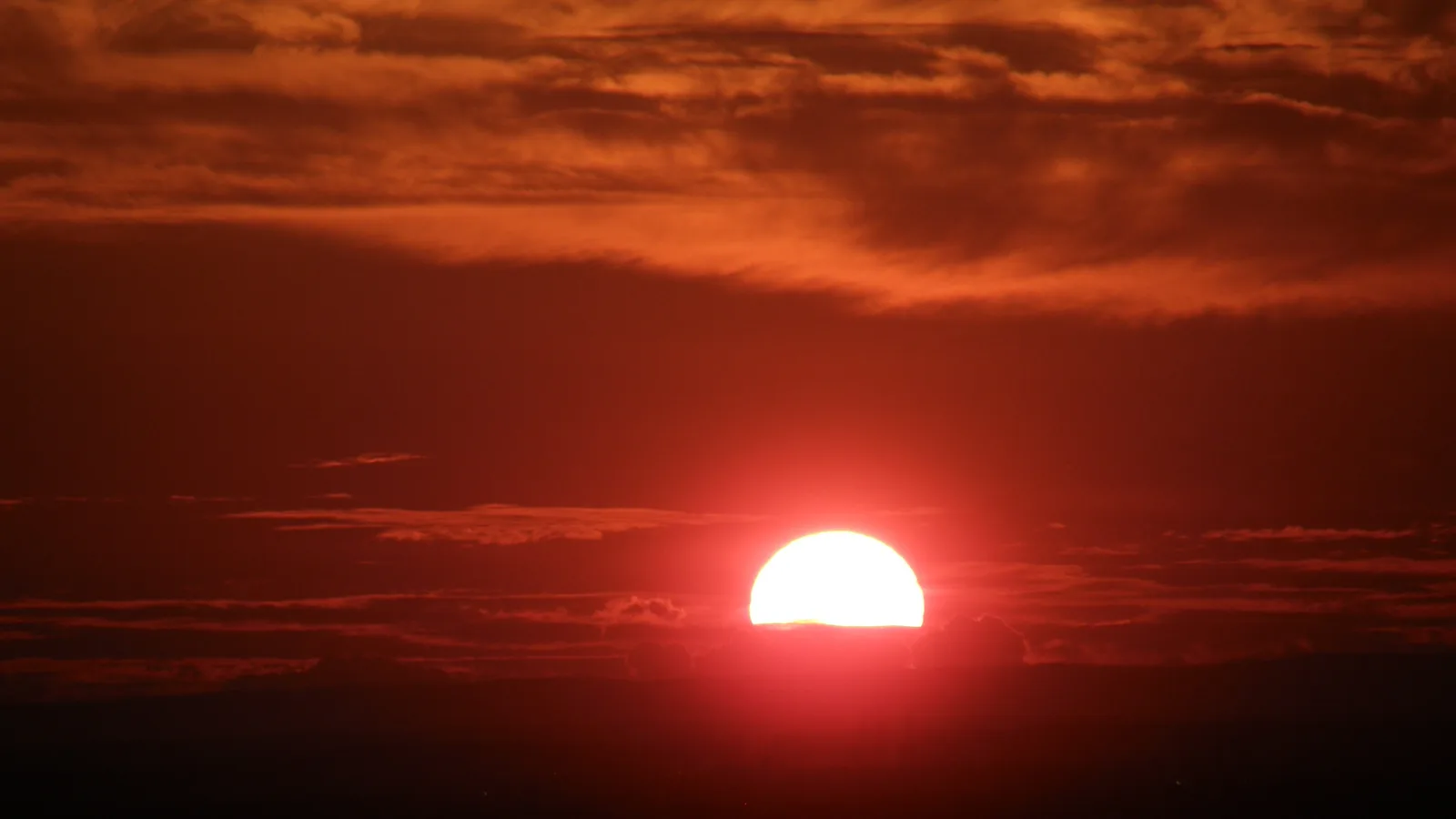 Tiefrote Sonne kurz über dem Horizont, davor dunkle Wolkenbänder in einem glühend orangefarbenen Himmel.