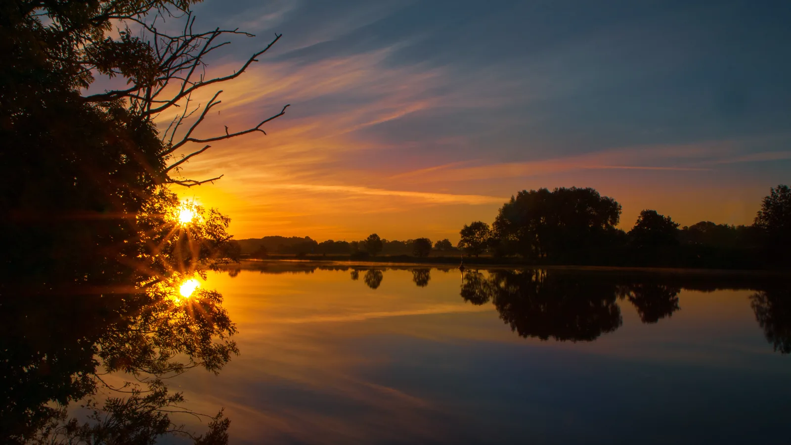 Sonnenaufgang über einem stillen See, die Sonne strahlt durch die Äste eines Baums am Ufer.