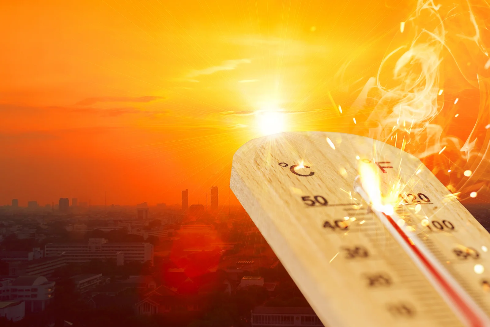 Glühendes Thermometer mit Funken vor oranger Sonnenglut über einer Stadt als Symbol für extreme Hitze.