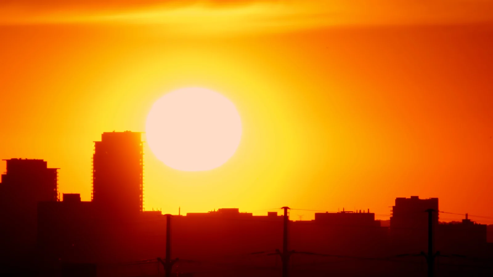 Glühend orangeroter Himmel mit der Sonne knapp über der Silhouette einer Stadt, die Gebäude zeichnen sich schwarz gegen das Licht ab.