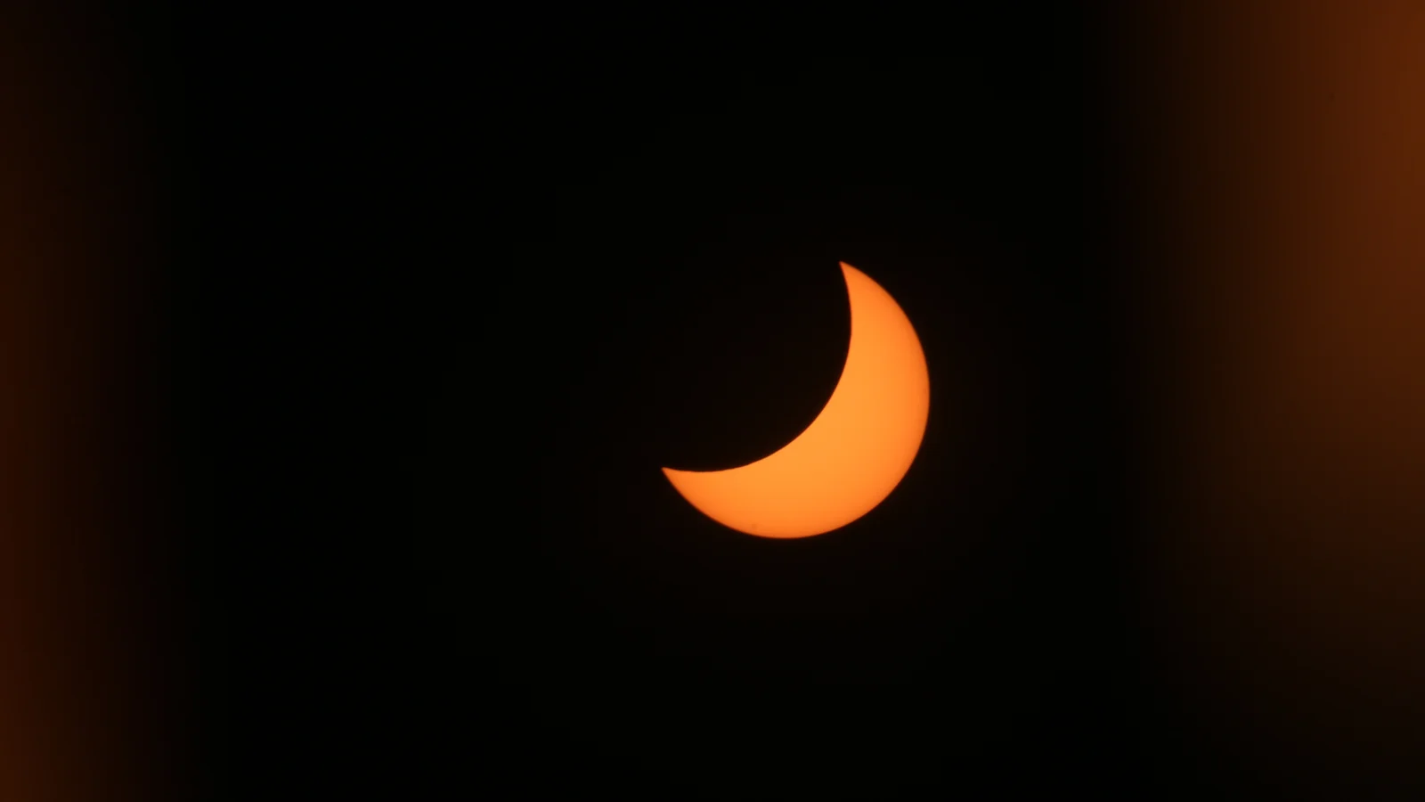 Teilweise verfinsterte Sonne als orange Sichel vor schwarzem Himmel, der Mond bedeckt den größten Teil der Sonne.