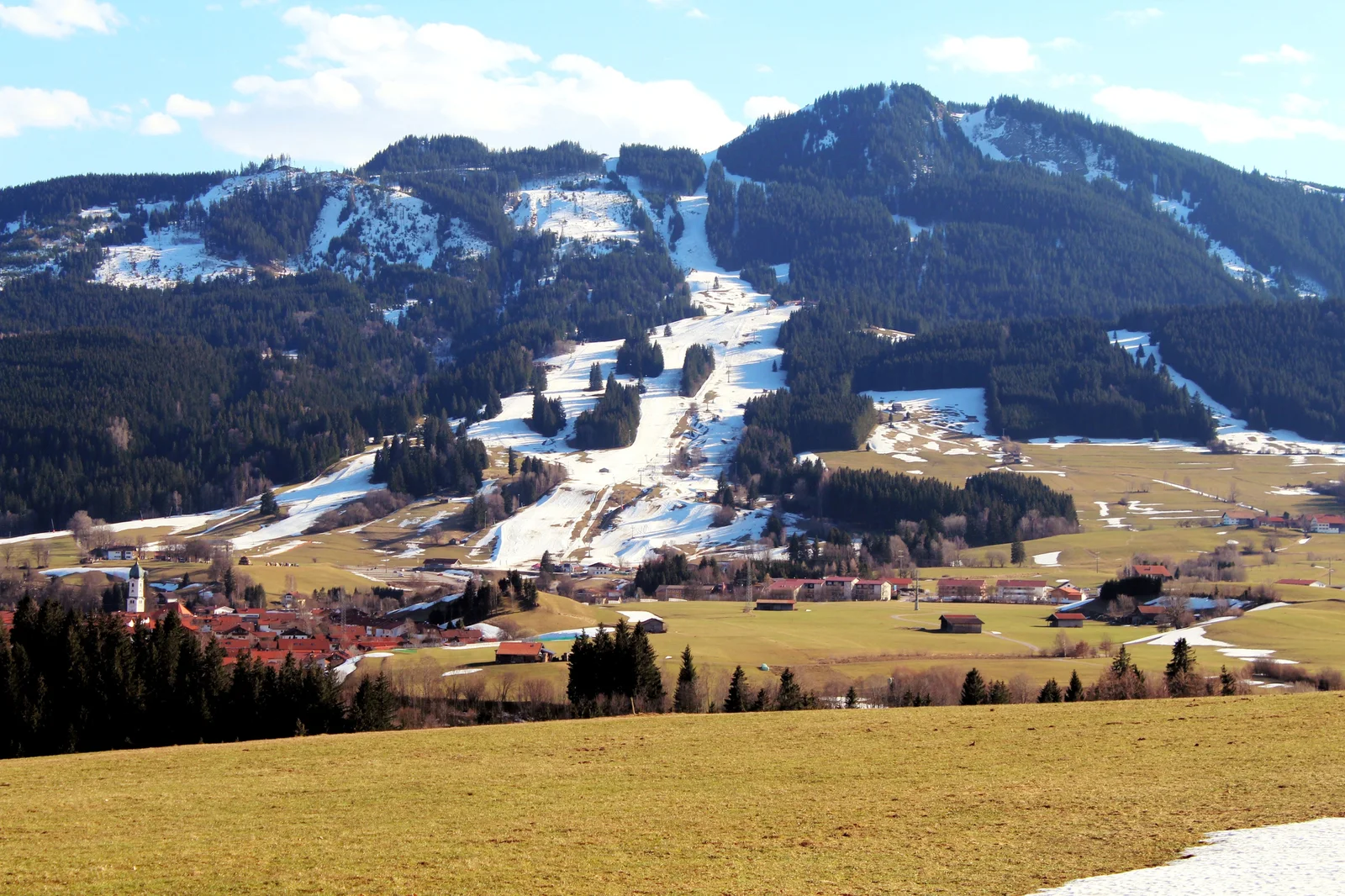 Skipisten aus Kunstschnee ziehen sich als weiße Bänder durch eine schneefreie Winterlandschaft.